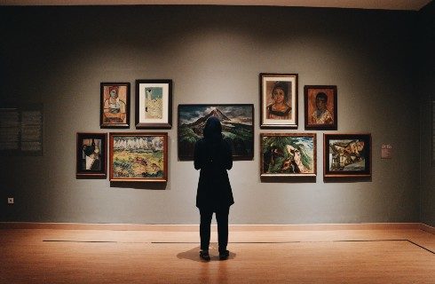 A person standing facing a lit up gallery wall with nine different paintings