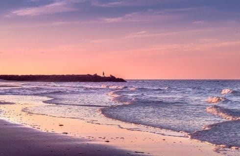 Wide open shore of a beach with soft waves and pink and purple sunrise skies