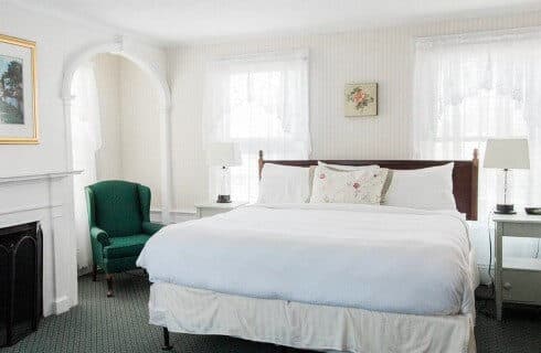 Bright bedroom with king bed, fireplace, green wingback chair and large windows with lace curtains