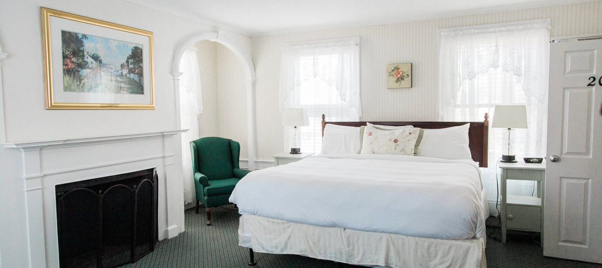 Bright bedroom with king bed, fireplace, green wingback chair and large windows with lace curtains