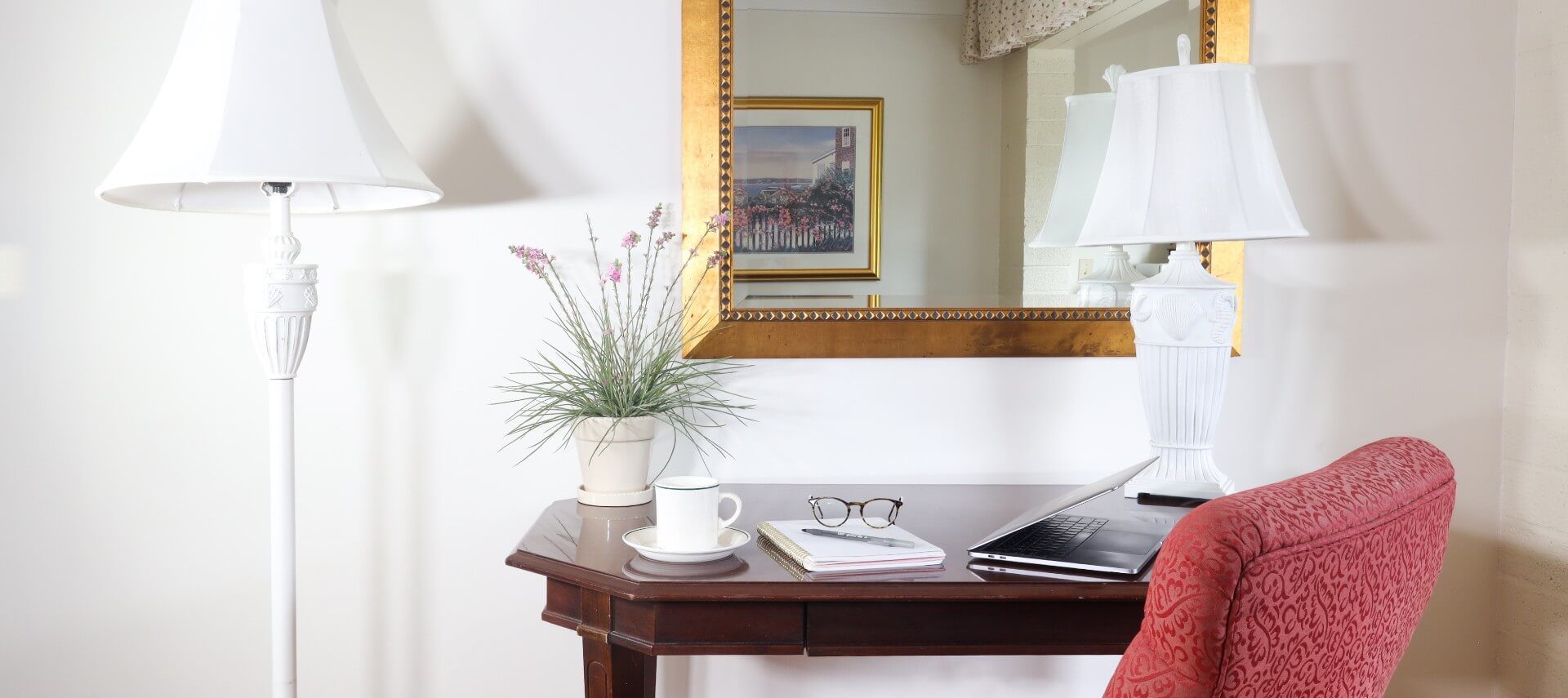 Writing desk with red chair, two white lamps, vase of flowers and gold framed mirror