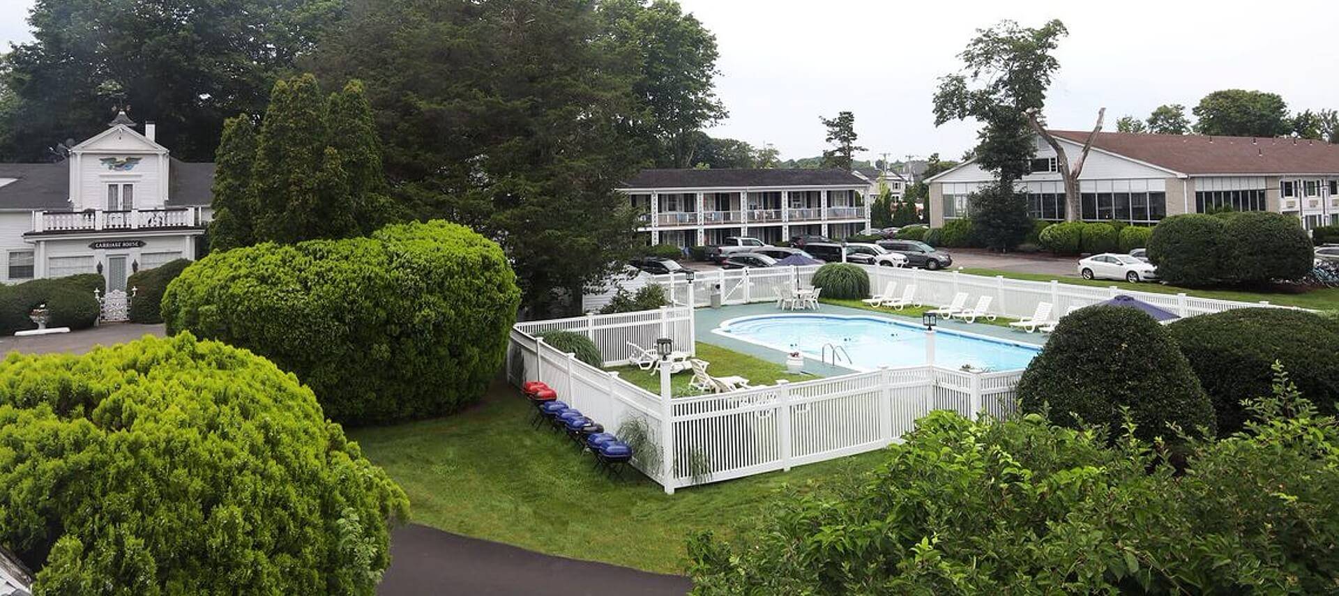 Outdoor pool with white fence surrounded by large trees, bushes and hotel buildings